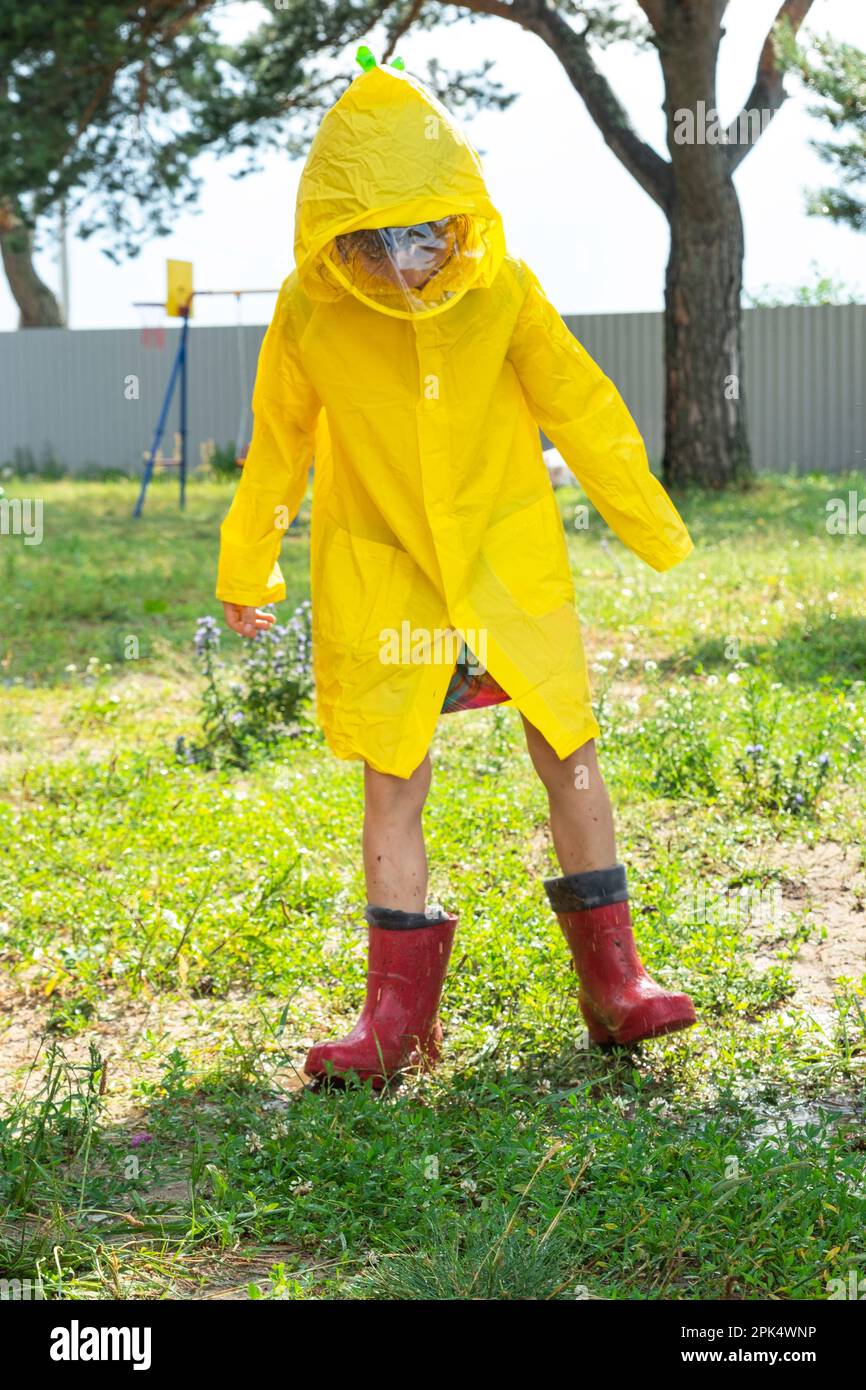 Una niña con botas de goma rojas y un impermeable amarillo corre a través  de charcos después de una lluvia en el pueblo. Tiempo de verano, libertad,  infancia Fotografía de stock -