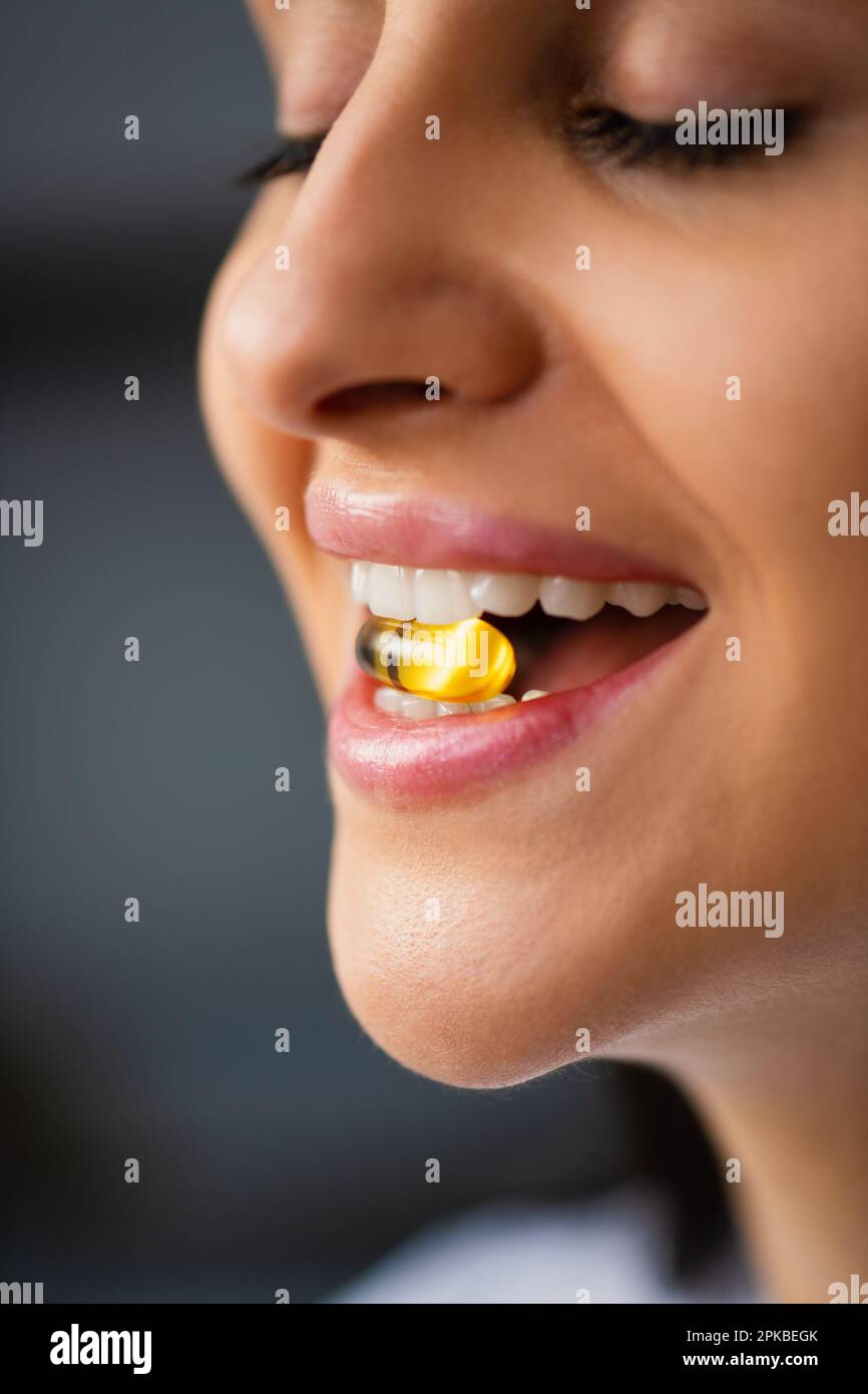 Mujer joven feliz multirracial que sostiene la boca amarilla de la píldora entre los dientes en aceite de pescado natural píldora de las vitaminas del omega 3 que se coloca por la ventana en casa Close-Up Foto de stock