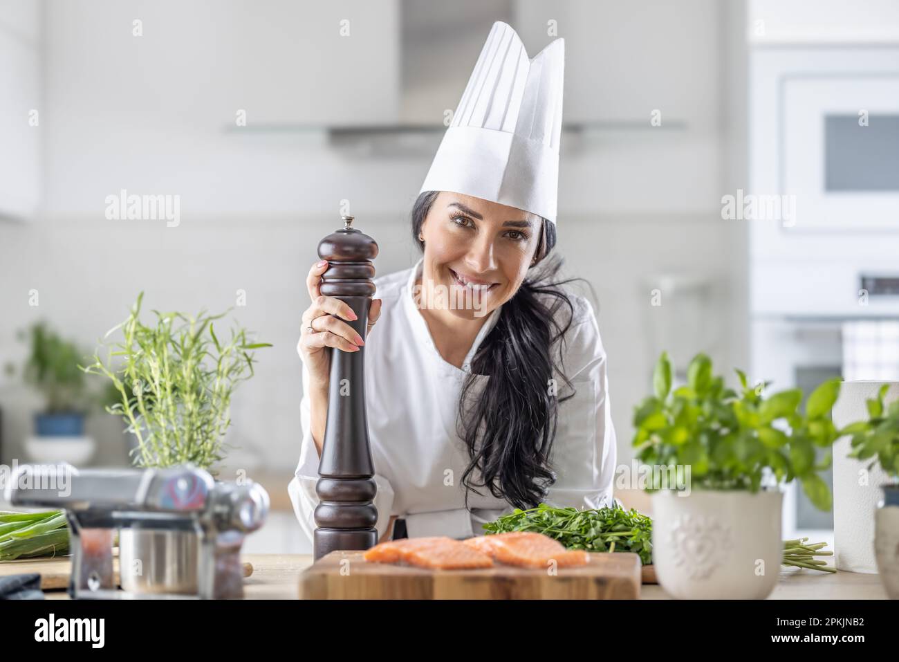 La chef femenina de toque blanche, sombrero blanco y uniforme sostiene un molinillo de pimienta durante la preparación de comidas de pescado y verduras. Foto de stock