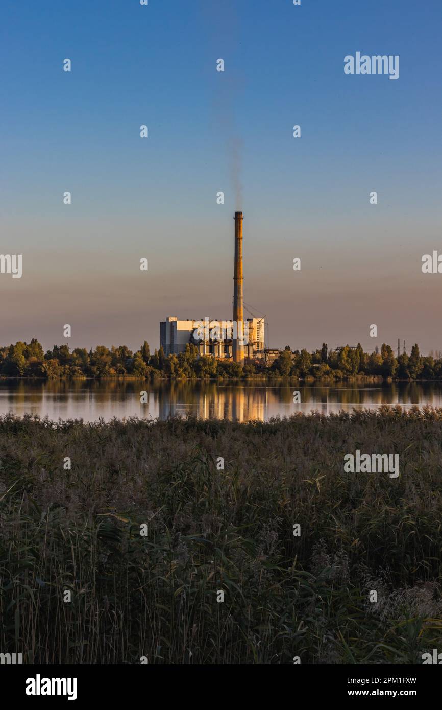 Una antigua planta de incineración de basura en la orilla del lago con chimeneas humeantes contra un cielo colorido. La generación de energía causa contaminación ambiental. Foto de stock