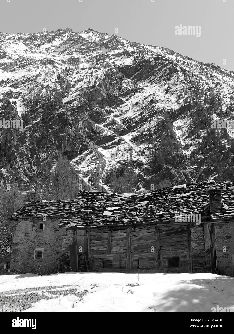 Edificio tradicional de madera bajo una montaña cubierta de nieve en Valnoney en el Parque Nacional Gran Paradiso, Valle de Aosta, Italia. Monocromo Foto de stock