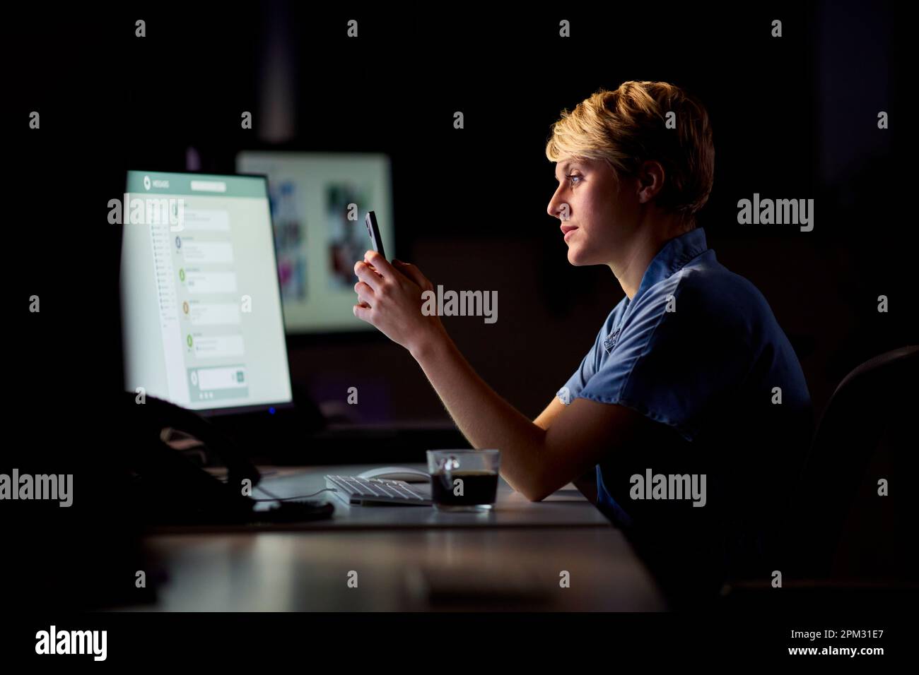 Empresaria que trabaja tarde en la oficina con la cara iluminada por la pantalla de la computadora usando el teléfono móvil Foto de stock