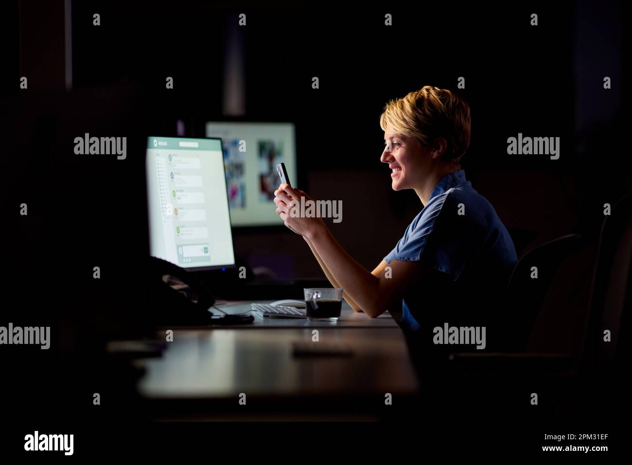 Empresaria que trabaja tarde en la oficina con la cara iluminada por la pantalla de la computadora usando el teléfono móvil Foto de stock