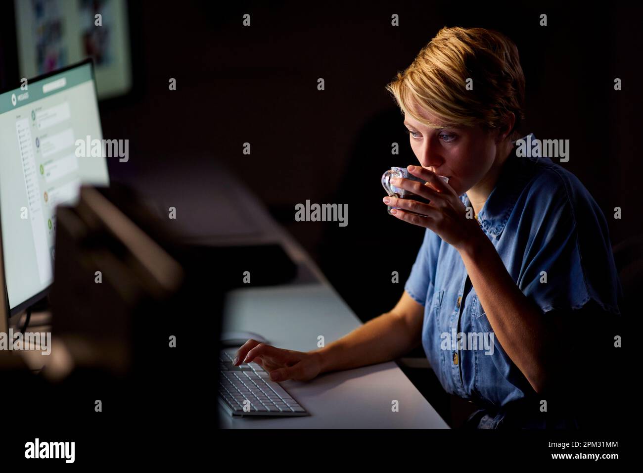 Empresaria que trabaja tarde en la oficina con la cara iluminada por la pantalla de la computadora que bebe café Foto de stock