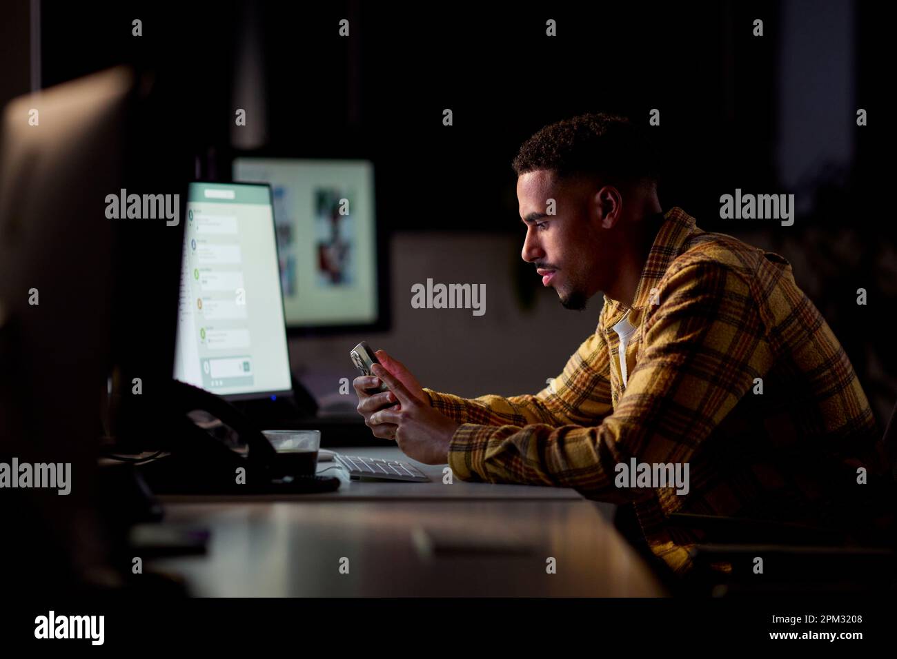 Hombre de negocios que trabaja tarde en la oficina con la cara iluminada por la pantalla de la computadora usando el teléfono móvil Foto de stock