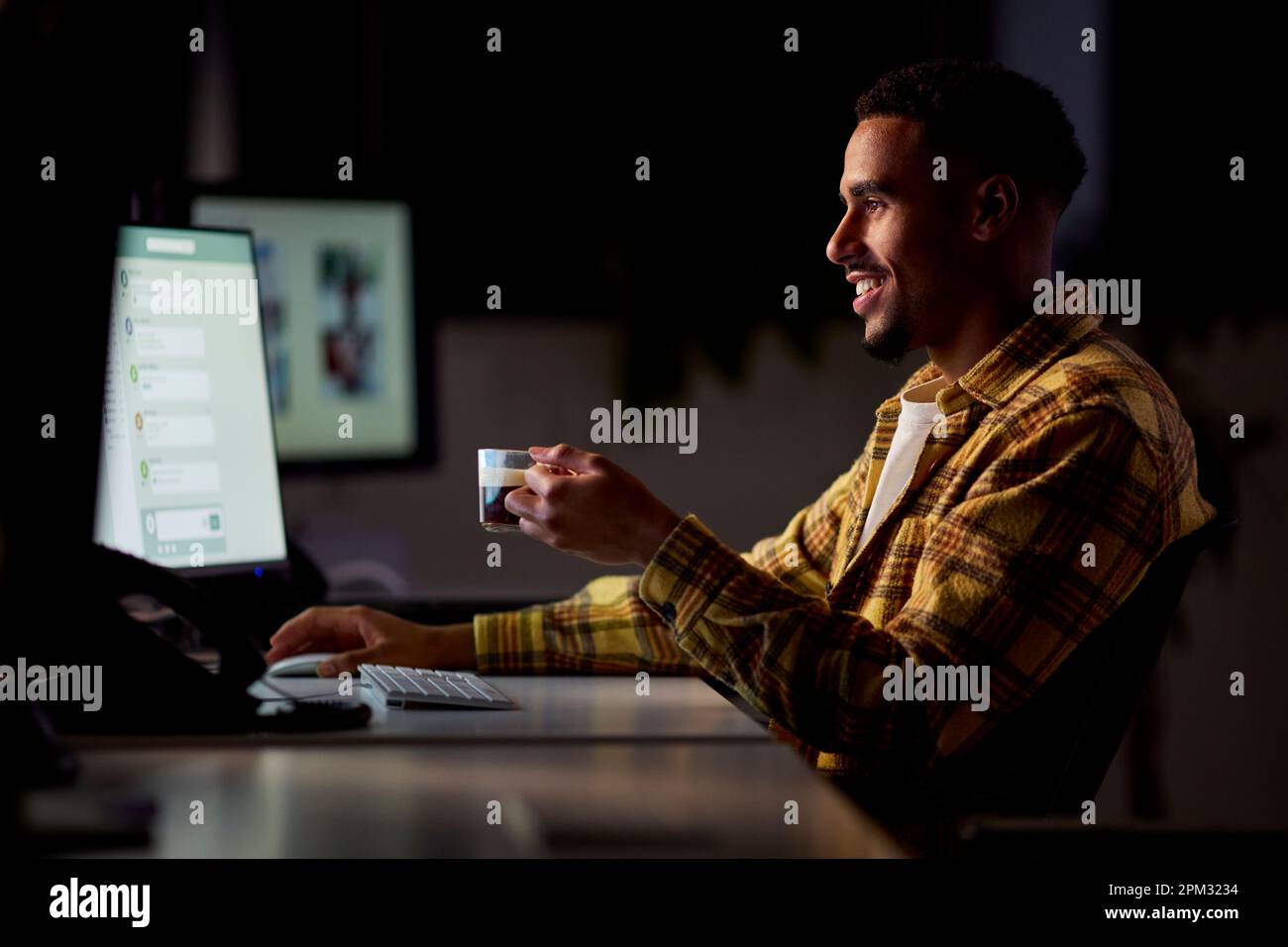 Hombre de negocios que trabaja tarde en la oficina con la cara iluminada por la pantalla de la computadora que bebe café Foto de stock