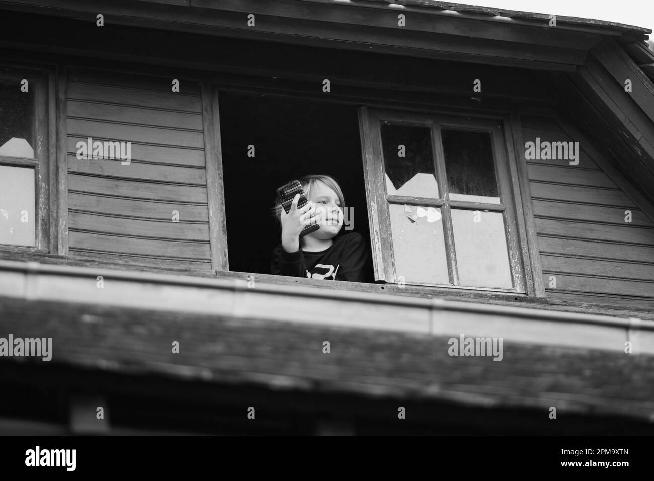 Un niño mira por la ventana de una casa de madera. Imagen en blanco y negro. Arresto domiciliario, descanso en el pueblo. Foto de stock