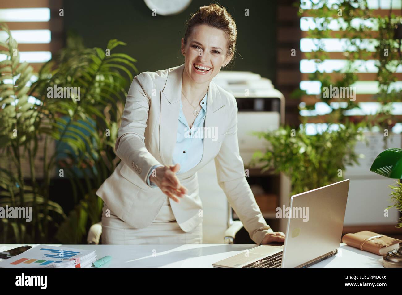 Retrato de mujer de negocios moderna feliz de 40 años en un traje de negocios ligero en la oficina verde moderna con el ordenador portátil dando la mano para un apretón de manos. Foto de stock