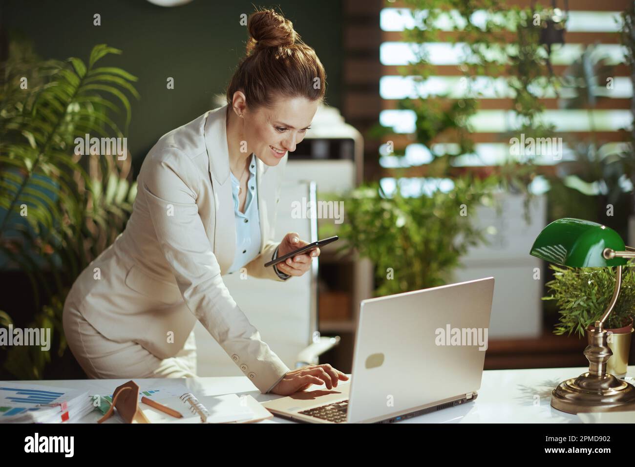 empleado moderno sonriente de la mujer de mediana edad en un traje ligero del negocio en la oficina verde moderna con el ordenador portátil usando la aplicación del smartphone. Foto de stock