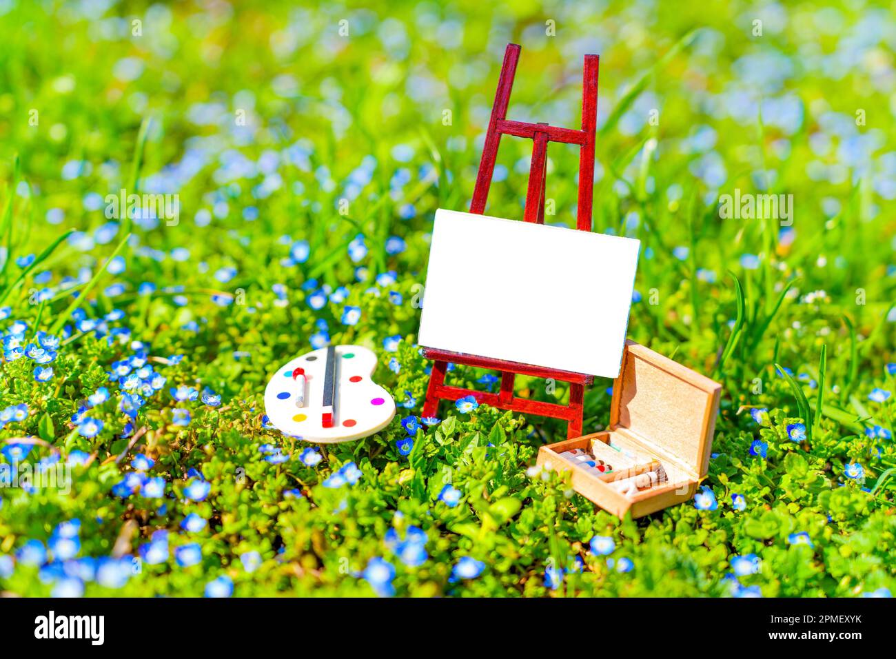 Lienzo pequeño en un pequeño soporte, paleta en miniatura y caja de madera con pinturas y pinceles en un exuberante campo en flor. Suministros de arte y concepto de equipo. Foto de stock