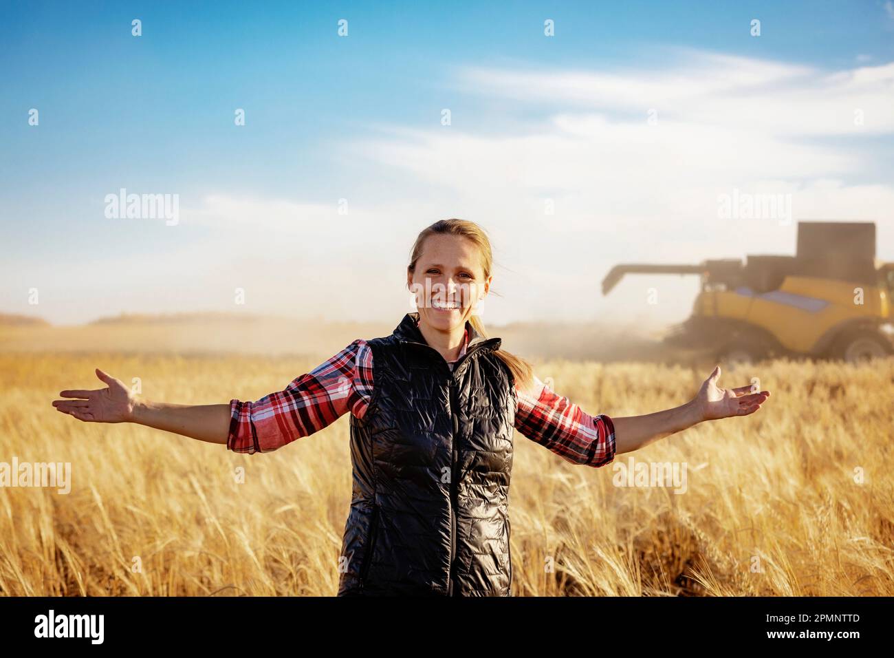 Retrato de una mujer de granja madura de pie en un campo de granos con los brazos extendidos, posando para la cámara durante la cosecha con una... Foto de stock