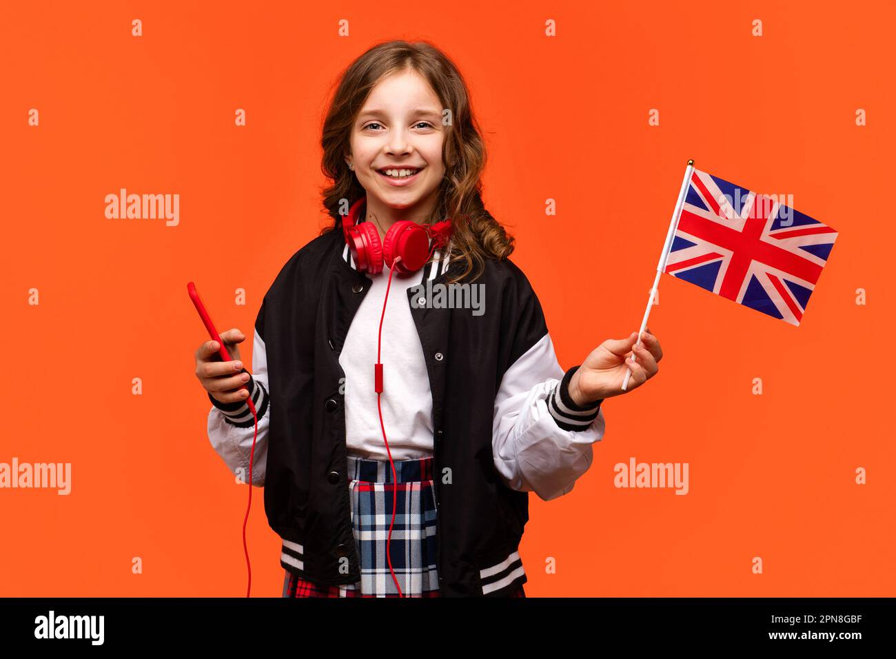 Adolescente en ropa casual y auriculares rojos con teléfono móvil. Chica de  la escuela sostiene la pequeña bandera del Reino Unido. Concepto de estudio  en el extranjero. Aprender inglés británico con nativo