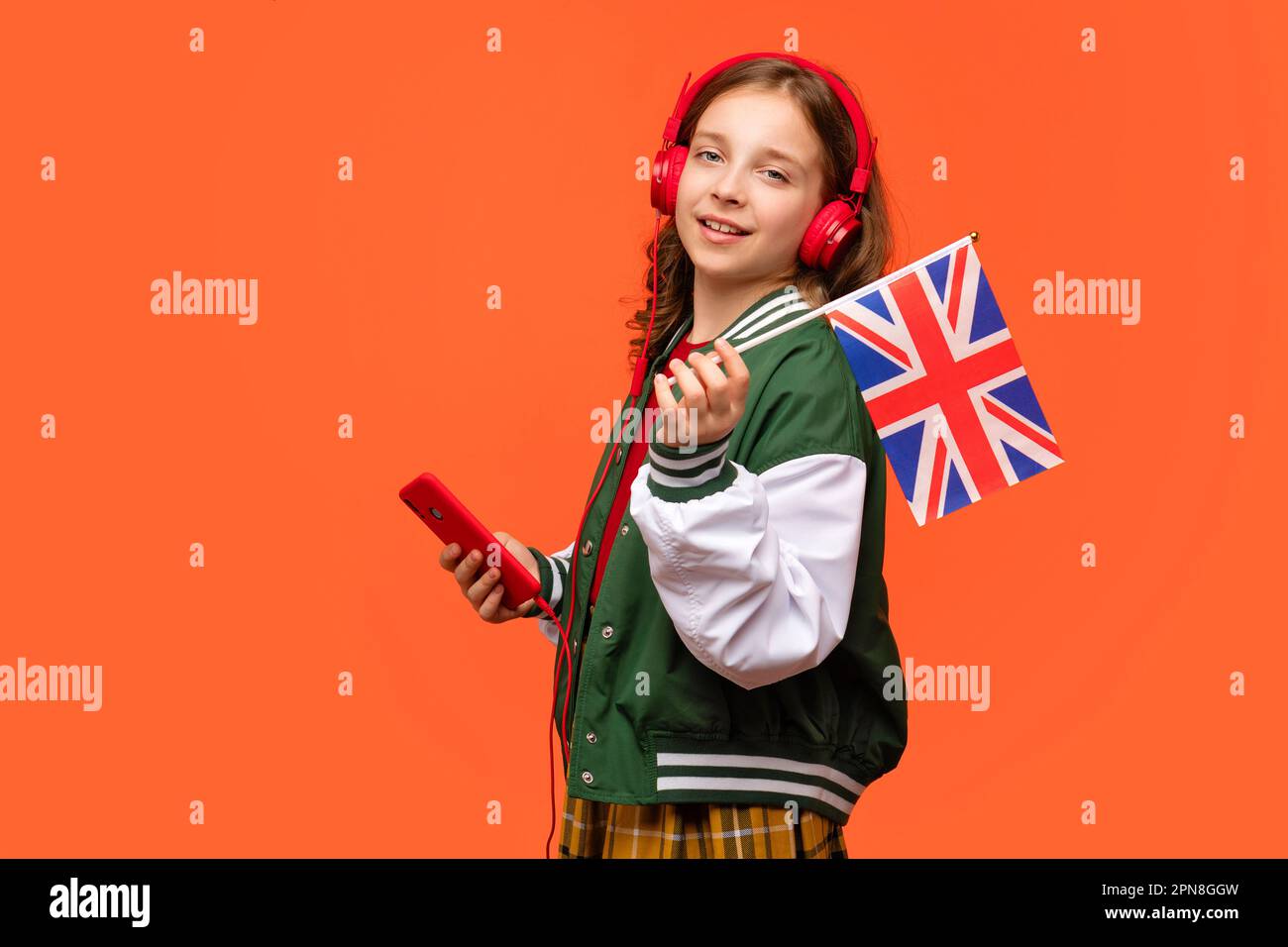 Chica de la escuela sostiene la pequeña bandera del Reino Unido. Adolescente ropa casual y auriculares rojos con teléfono móvil. Cursos de idiomas online. Aprender inglés británico. Lista Fotografía de -