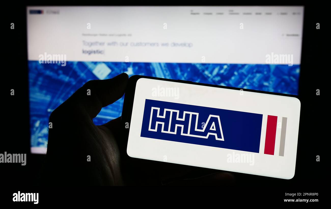 Persona que sostiene el teléfono inteligente con el logotipo de la empresa Hamburger Hafen und Logistik AG (HHLA) en la pantalla frente al sitio web. Enfoque en la pantalla del teléfono. Foto de stock