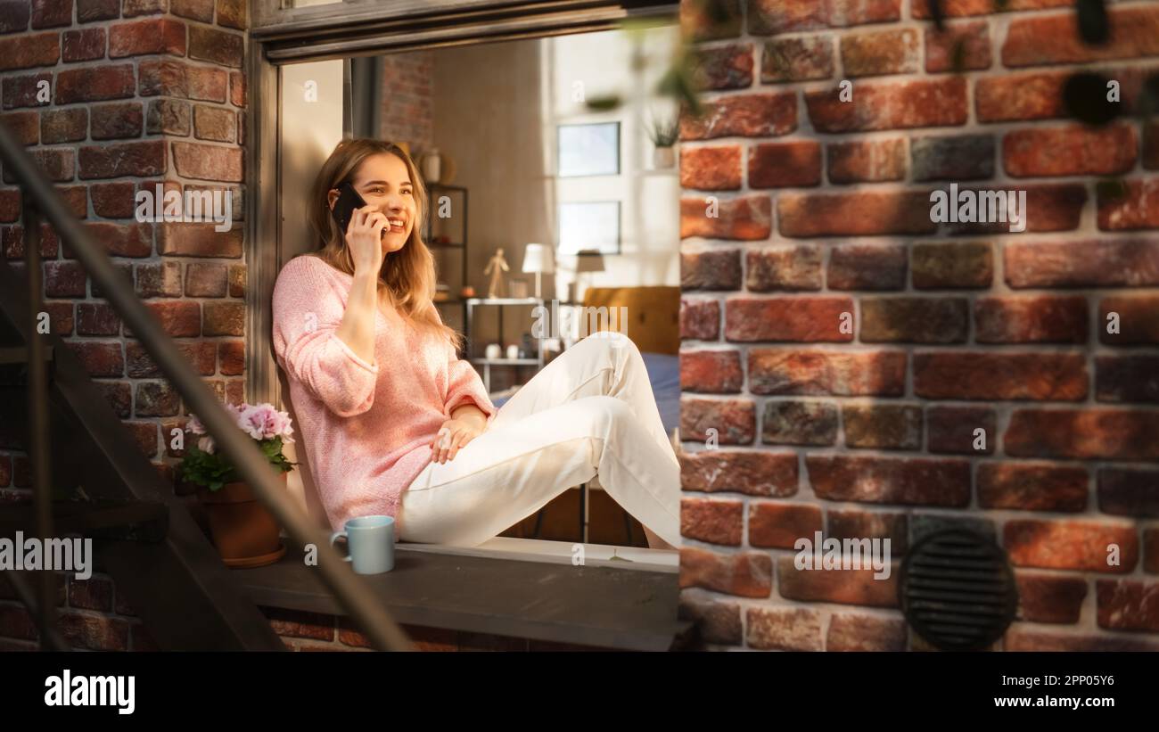 Hermosa joven mujer que tiene una llamada en su teléfono inteligente mientras se sienta en su alféizar de ventana. Mujer expresiva hablando por teléfono, hablando. Foto de stock