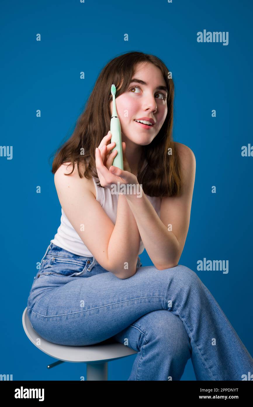 Feliz adolescente sosteniendo cepillo de dientes eléctrico sentado | Fondo azul Foto de stock