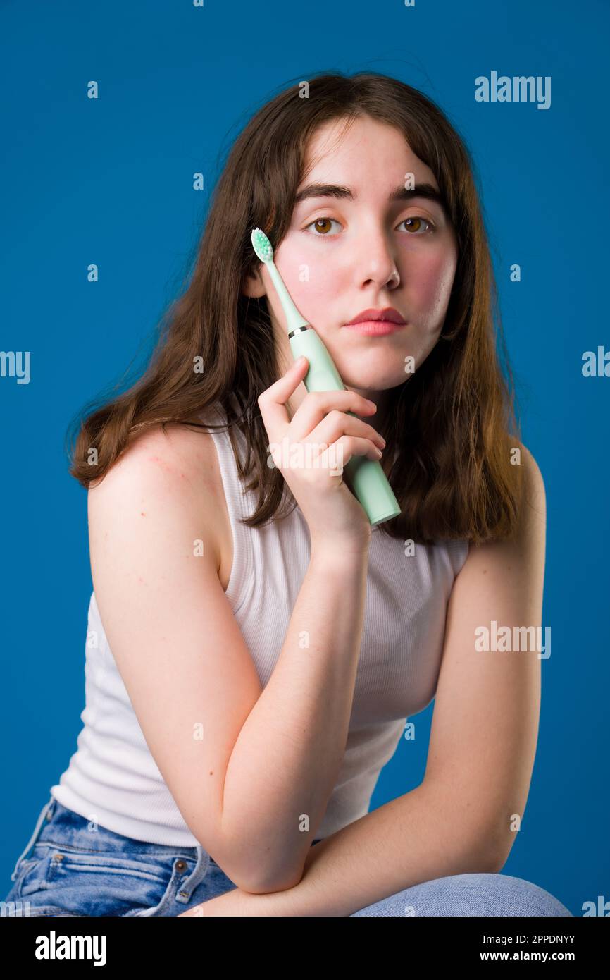 Feliz adolescente sosteniendo cepillo de dientes eléctrico sentado | Fondo azul Foto de stock