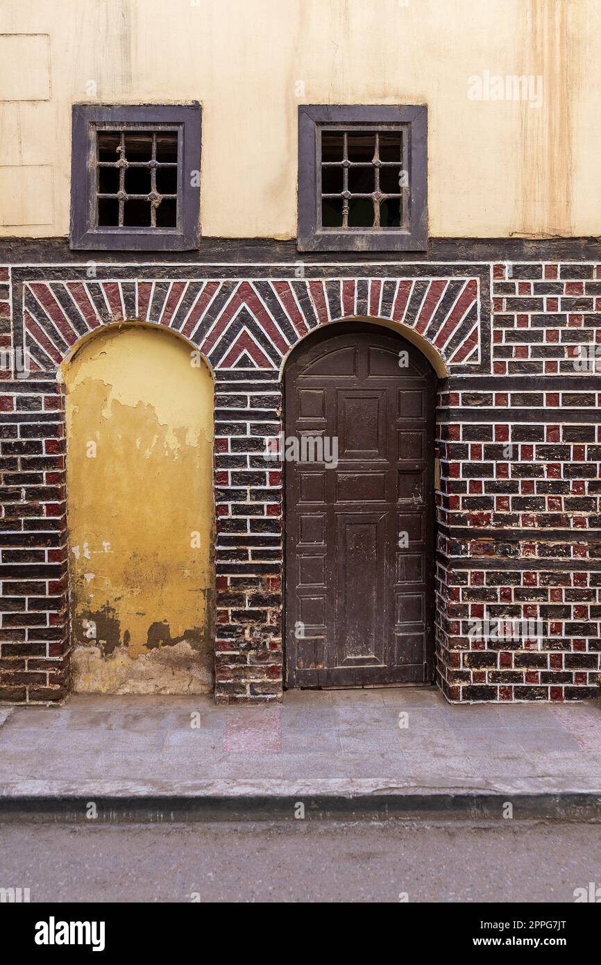 Puerta de arco bloqueada, puerta de madera y pequeñas ventanas con barras oxidadas en la pared de ladrillos negros y rojos Foto de stock