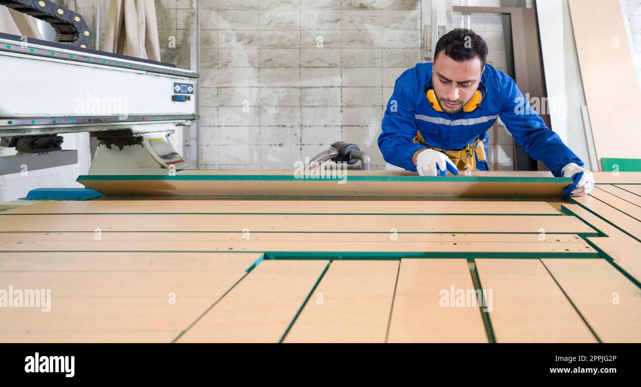 Técnico joven en mono mecánico azul, orejeras y cinturones de herramientas amarillos cargando madera contrachapada en la mesa de trabajo. Atmósfera de la línea de producción en una fábrica de muebles de madera. Foto de stock