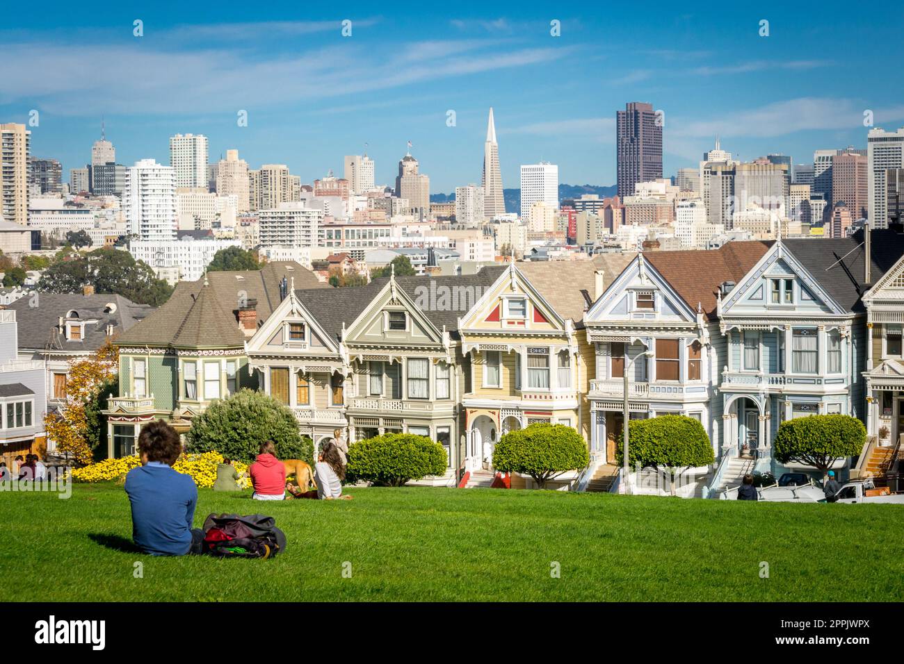 Vista en las casas victorianas pintadas de las damas de San Francisco con paisaje urbano y horizonte en el fondo en un cielo azul. Gente sentada delante Foto de stock