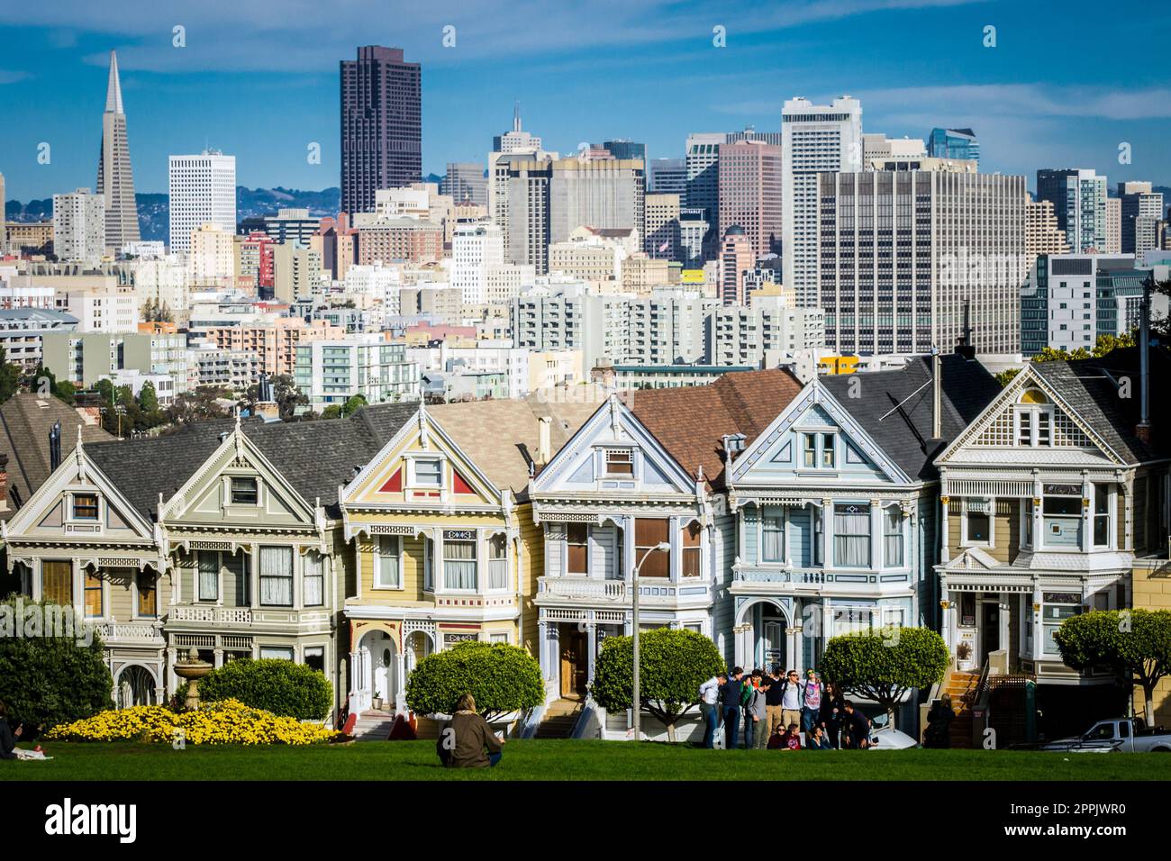 Vista en las casas victorianas pintadas de las damas de San Francisco con paisaje urbano y horizonte en el fondo en un cielo azul. Gente sentada delante Foto de stock
