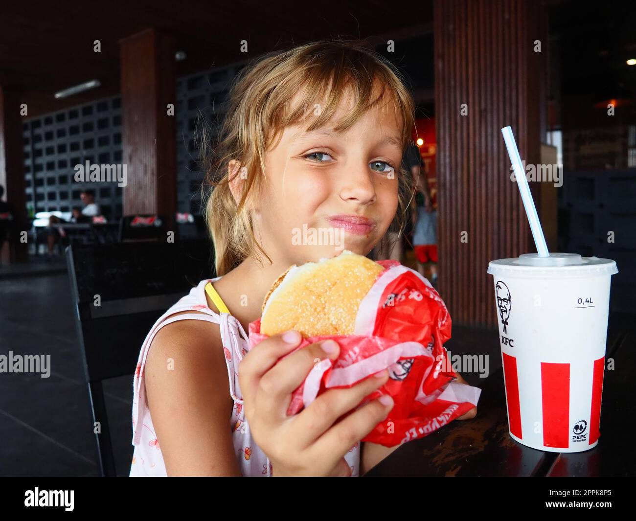 Anapa Rusia 23 agosto 2021 Una hermosa chica de 7 años está comiendo en el restaurante KFC disfrutando de la comida y sonriendo. Niño caucásico sosteniendo una hamburguesa. Cola en una taza de cartón con tapa y paja Foto de stock
