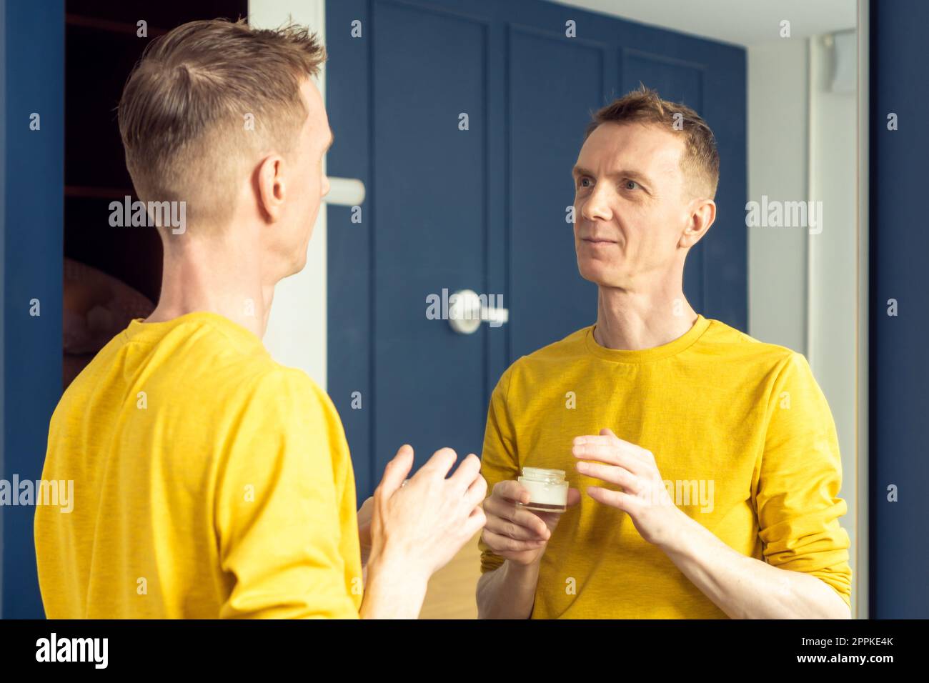 El hombre maduro feliz sostiene el tarro de crema hidratante y aplica sobre la piel clara de la cara. Retrato masculino en el espejo de la habitación. Foto de stock