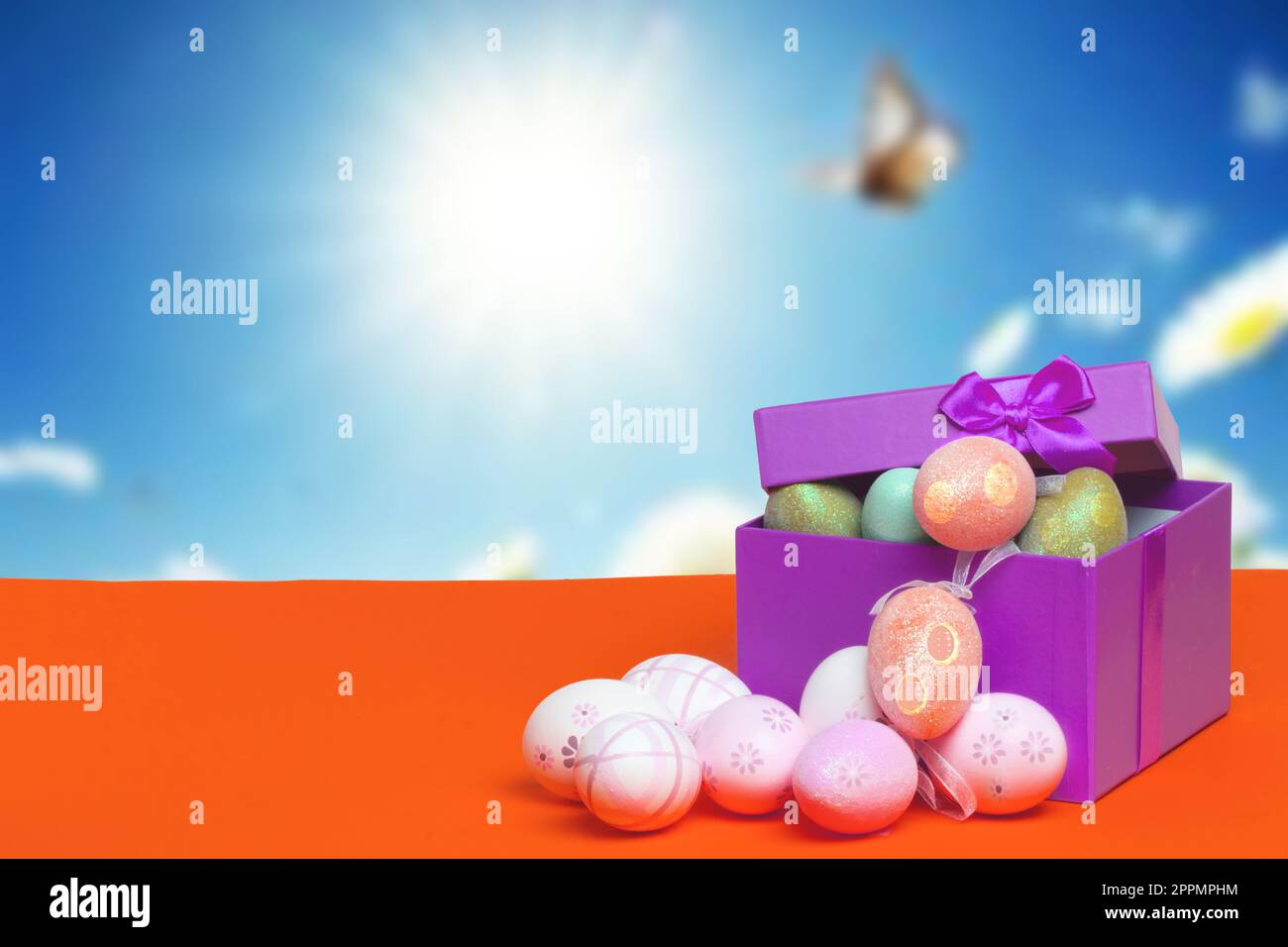 Plantilla de tarjeta de felicitación de feliz Pascua. Cierre para arriba de los huevos de Pascua coloridos y una caja de regalo abierta con los huevos en la mesa naranja contra el fondo soleado borroso abstracto del cielo. Copiar espacio para el diseño. Foto de stock