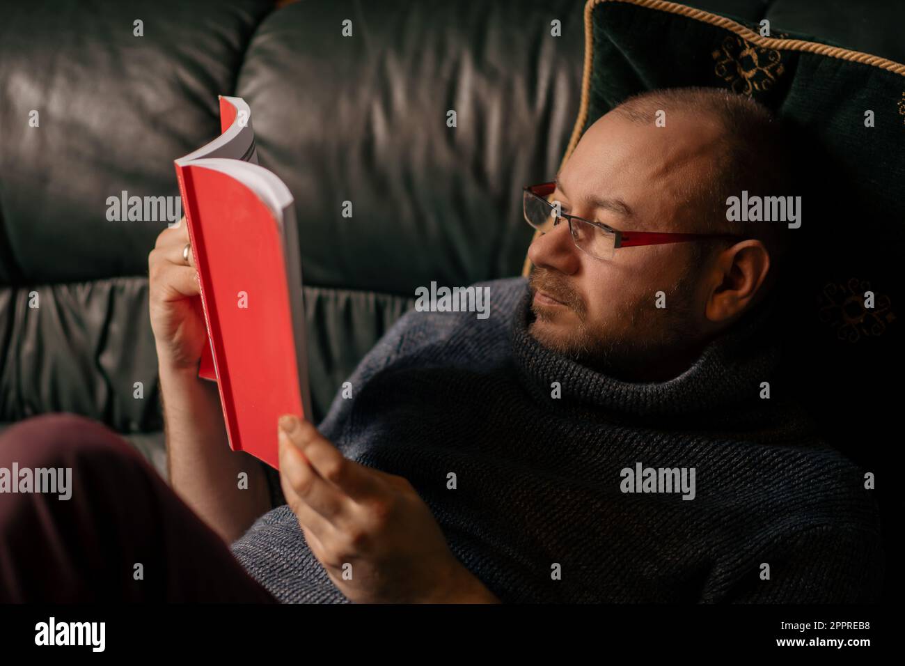 Un hombre con gafas está leyendo un libro mientras está acostado en el  sofá. Hombre de mediana edad de apariencia ordinaria en ropa casual en  casa. Leer, aprender Fotografía de stock -