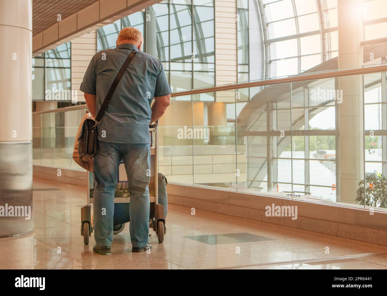 Una figura de cuerpo entero de un hombre en jeans azules y una camisa, con un carrito de equipaje, en el fondo del interior del aeropuerto, una copia del espacio a la derecha Foto de stock