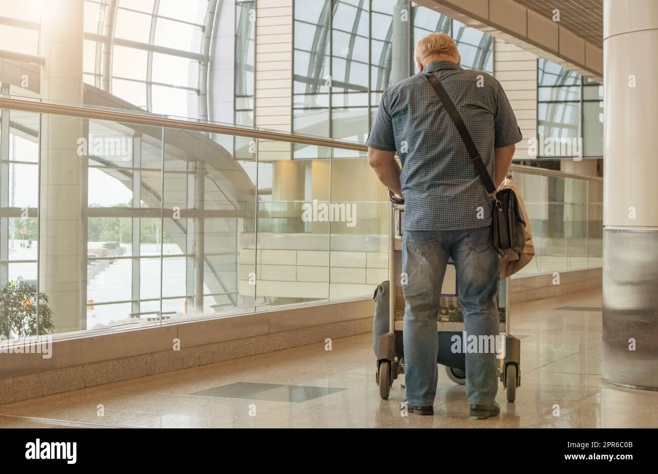 Una figura de cuerpo entero de un hombre en jeans azules y una camisa, con un carrito de equipaje, en el fondo del interior del aeropuerto, una copia del espacio a la izquierda Foto de stock
