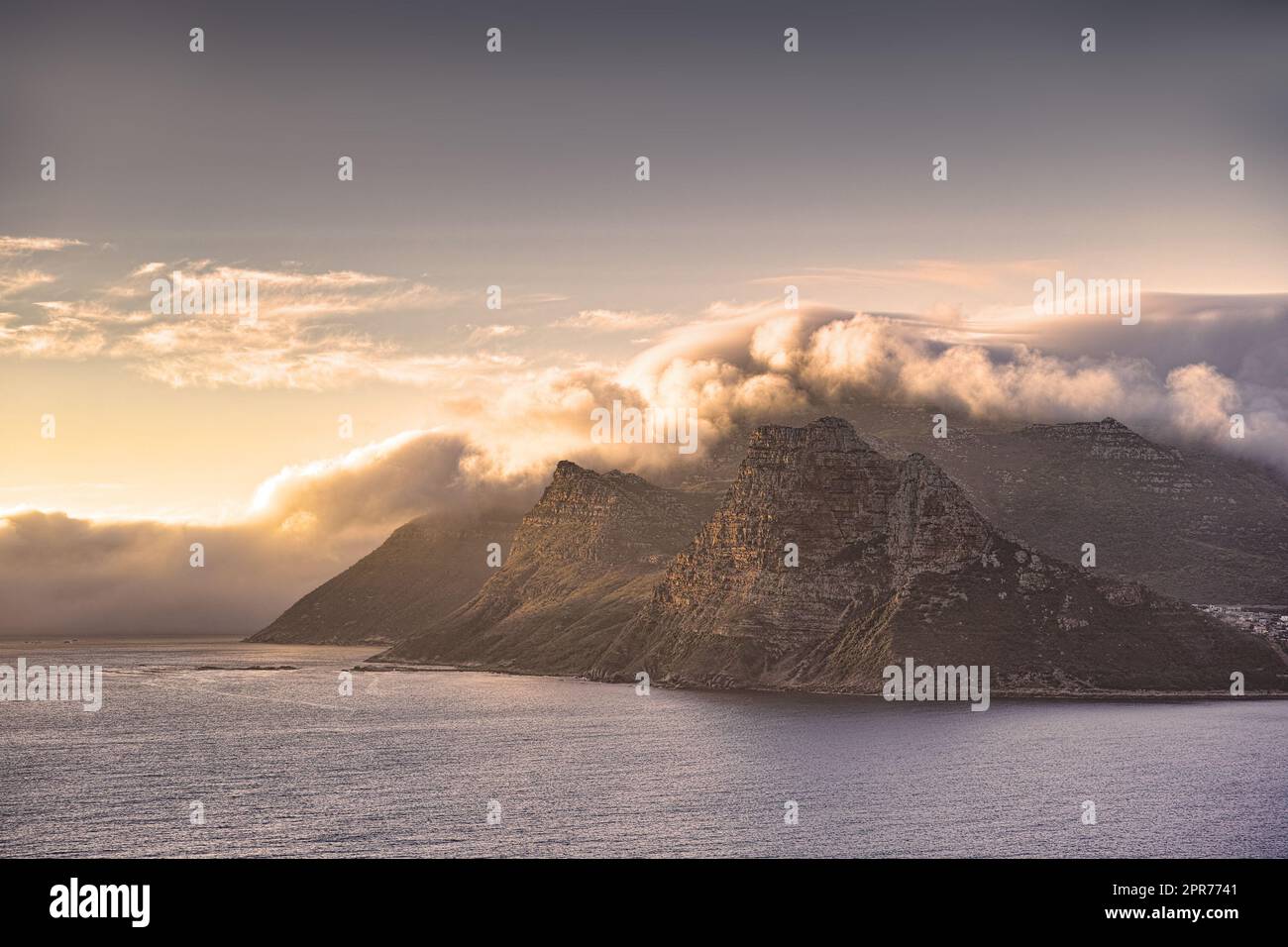 Panorama de una costa de montaña con una puesta de sol nublada en Sudáfrica. Paisaje escénico de suaves nubes blancas que cubren la Montaña de la Mesa al atardecer cerca de un mar tranquilo y pacífico en Hout Bay cerca de Ciudad del Cabo. Foto de stock