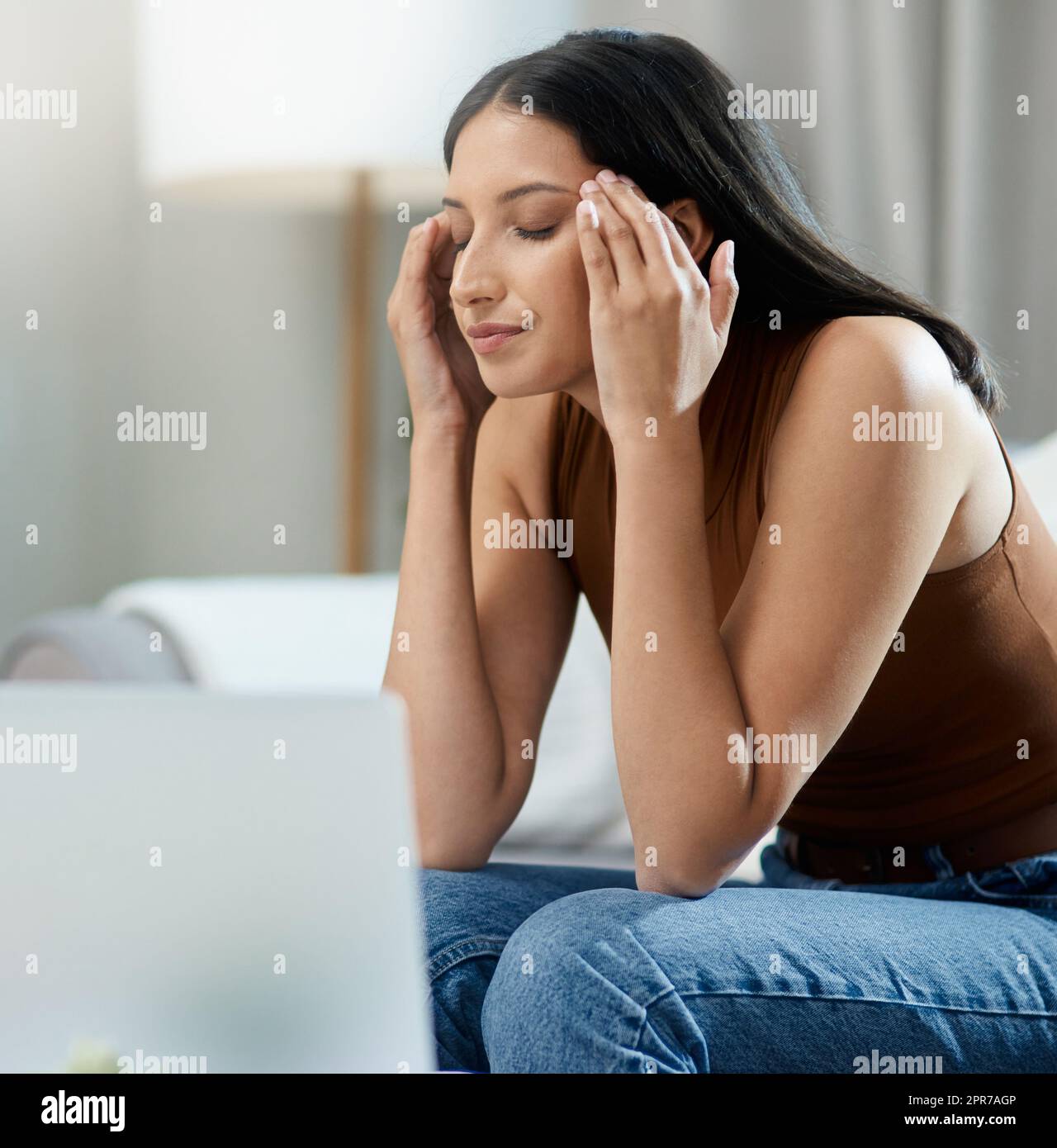 Mi cabeza me está matando. Una atractiva mujer joven sentada sola en su sala de estar y sintiéndose estresada mientras usa su ordenador portátil. Foto de stock