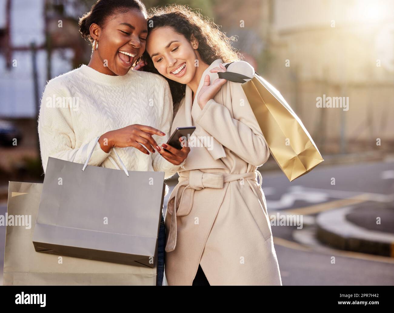 Compra lo mejor por mucho menos. Dos mujeres jóvenes que usan un smartphone mientras hacen compras con un fondo urbano. Foto de stock