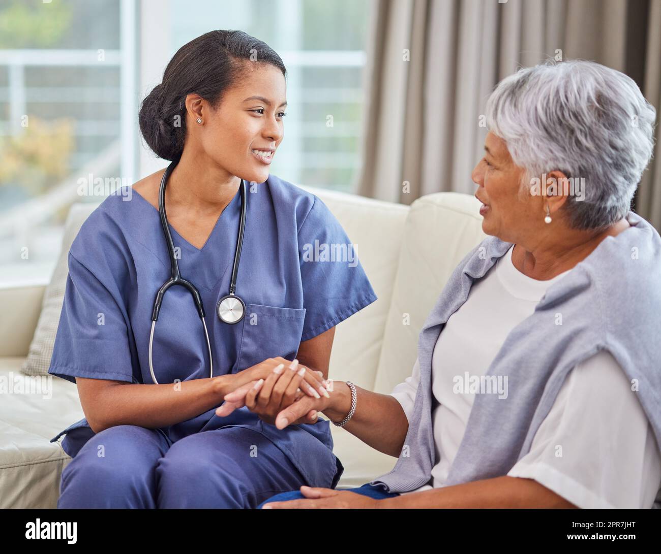 Una mujer hispana de la tercera edad en un cómodo sofá y su enfermera en el hogar de la tercera edad. Raza mixta joven enfermera y su paciente hablando en el salón Foto de stock
