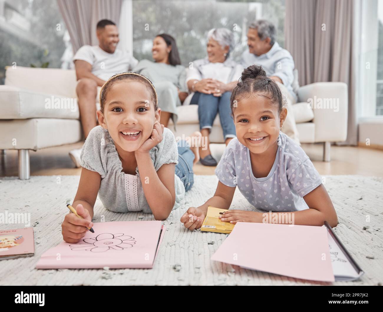 Dos hermanas hermanas hermanas lindas de raza mixta dibujando y coloreando en la sala de estar con sus padres y abuelos en el fondo. Niños despreocupados jugando mientras mamá, papá, abuelita y abuelo miran Foto de stock