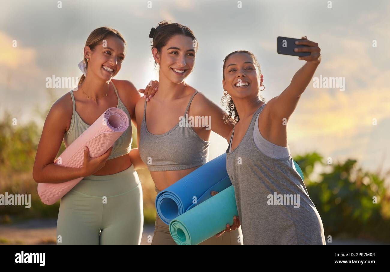 Hermosas mujeres de yoga que usan un teléfono móvil para tomar selfies mientras sostienen esteras de yoga en la práctica al aire libre en la naturaleza remota. Diverso grupo de jóvenes amigos activos sonrientes de pie juntos. Tres personas felices Foto de stock