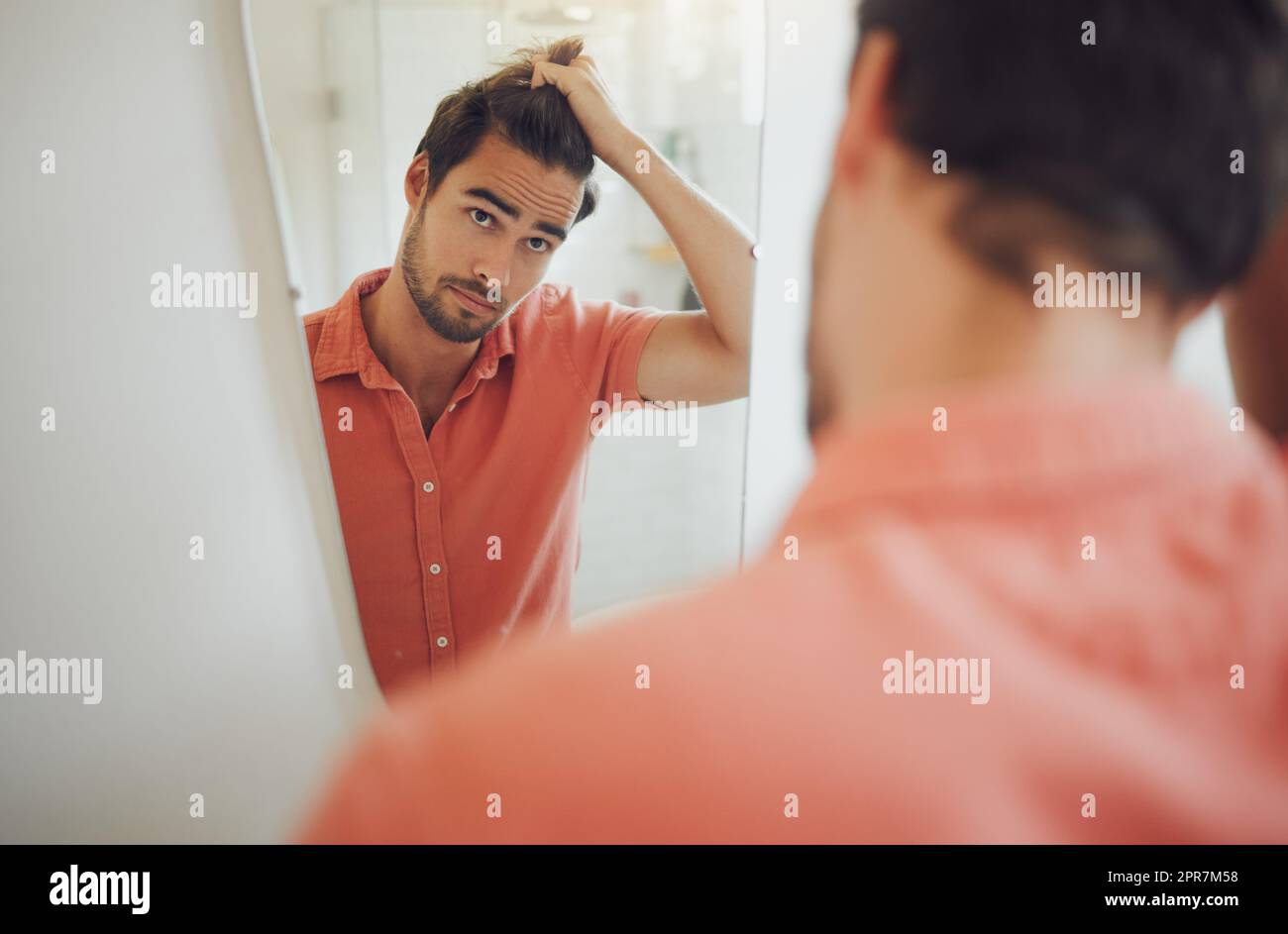 Guapo joven caucásico tocando su cabello y mirando en el espejo del baño. Hombre tirando de su pelo y pensando en conseguir un corte de pelo. Hombre preocupado por la caspa, el retroceso de la línea del cabello o la pérdida del cabello Foto de stock