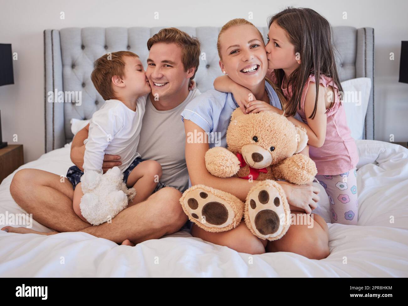 Los niños que aman compartiendo el momento tierno con los padres mientras besan suavemente a mamá y papá en la mejilla. Felices padres caucásicos pasando tiempo con su hija pequeña y su hijo en casa por la mañana Foto de stock