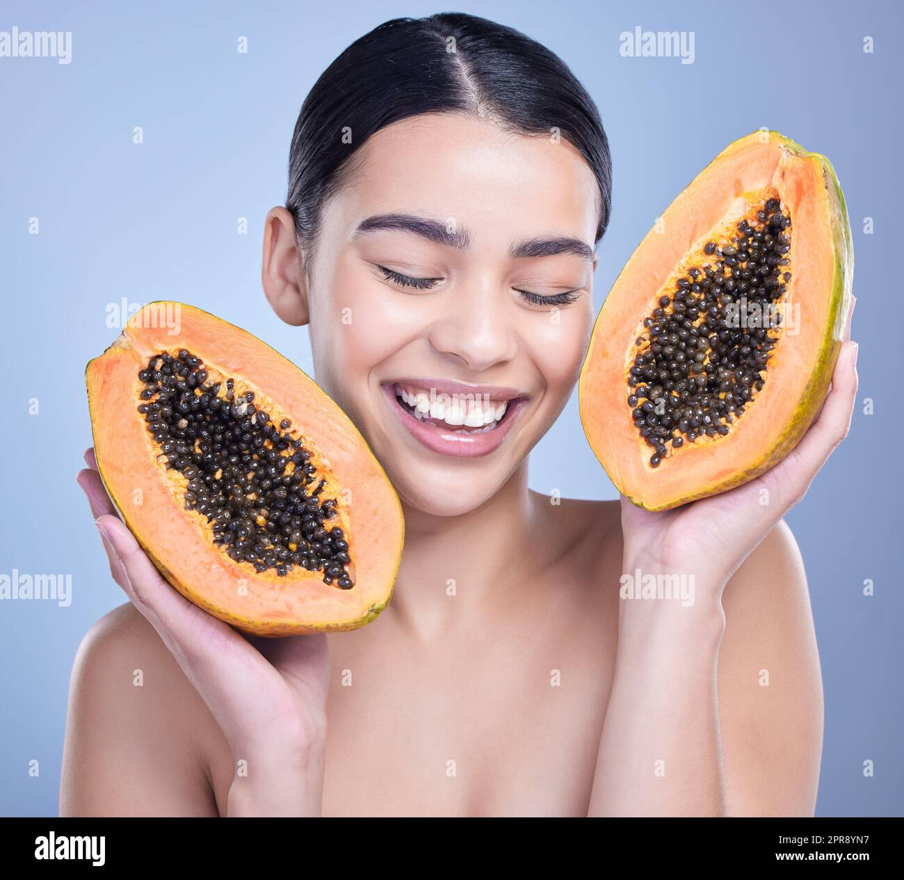 Una mujer de raza mixta sonriente feliz sosteniendo una papaya. Modelo hispano que promueve los beneficios de una dieta saludable en la piel contra un fondo azul de copyspace Foto de stock