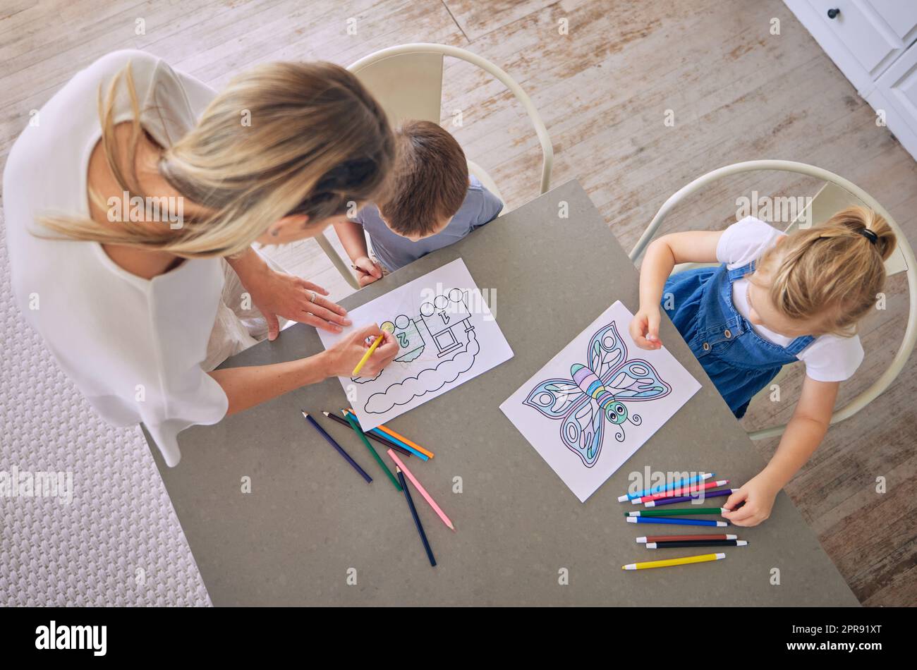 Encima de la foto de la niña y el niño sentado en la mesa con lápices de colores e imágenes mientras que colorean con la ayuda de la mamá. Madre caucásica con dos hijos disfrutando del pasatiempo educativo y siendo creativo Foto de stock