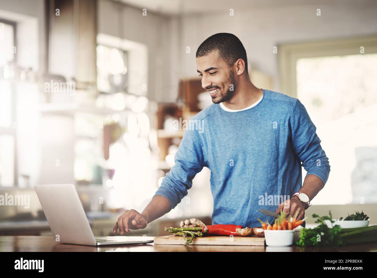 Hombre casual joven en la cocina en una computadora portátil mientras prepara una comida saludable en casa. Feliz hombre sonriente navegando y aprendiendo en la computadora sobre cómo cocinar. Chico solo comprobando recetas en línea en la web. Foto de stock