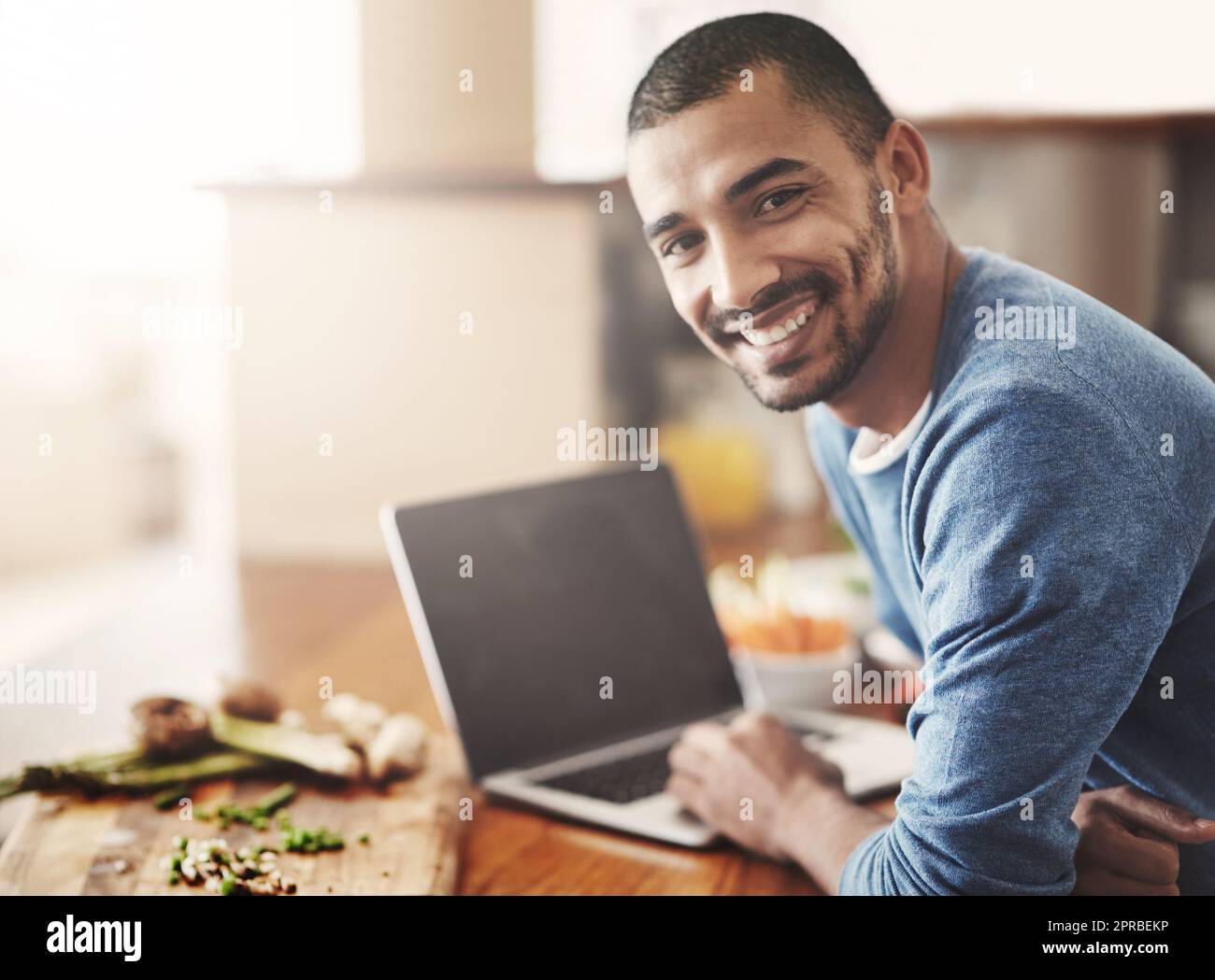 Hombre joven en una computadora portátil mientras prepara una comida saludable en casa. Retrato feliz sonriente masculino navegando y aprendiendo en la computadora en la cocina sobre cómo cocinar. Chico solo comprobando recetas en línea en la web Foto de stock