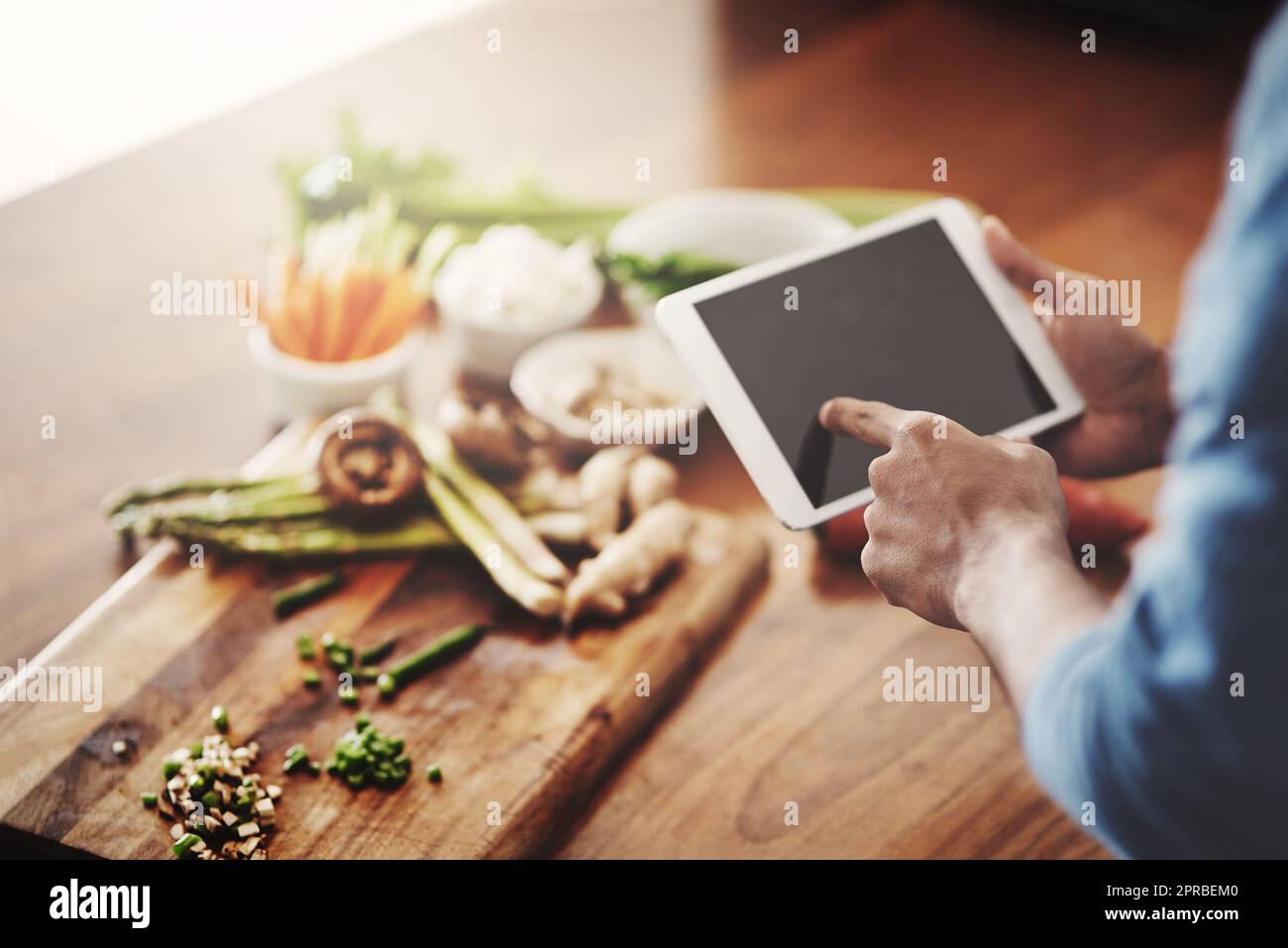 Primer plano de las manos del hombre sosteniendo una tableta para investigar recetas saludables, ver videos tutoriales de cocina y desplazarse en línea para ideas de comidas mientras prepara la cena, el almuerzo o el desayuno. Hombre navegando en la aplicación Foto de stock