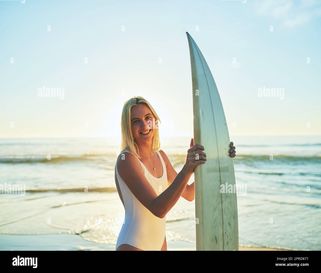 La vida es simplemente mejor cuando usted practica surf. Retrato recortado de una atractiva mujer joven de pie en traje de baño sosteniendo una tabla de surf en la playa. Foto de stock