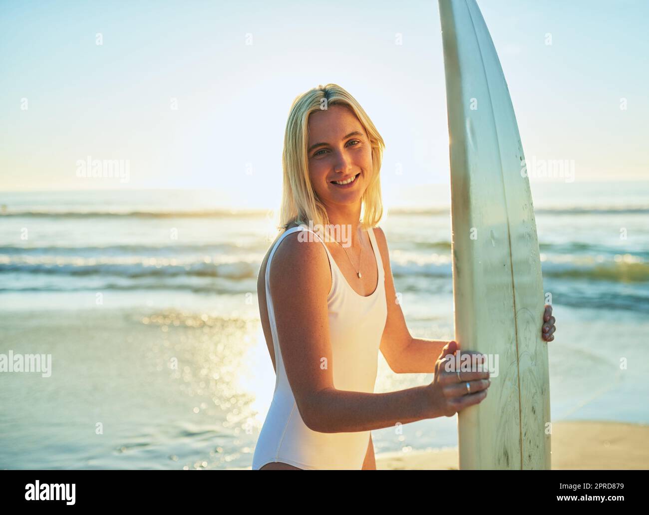 En mi más feliz cuando una tabla de surf está entre mis manos. Retrato recortado de una mujer joven atractiva de pie en un traje de baño sosteniendo una tabla de surf en la playa. Foto de stock