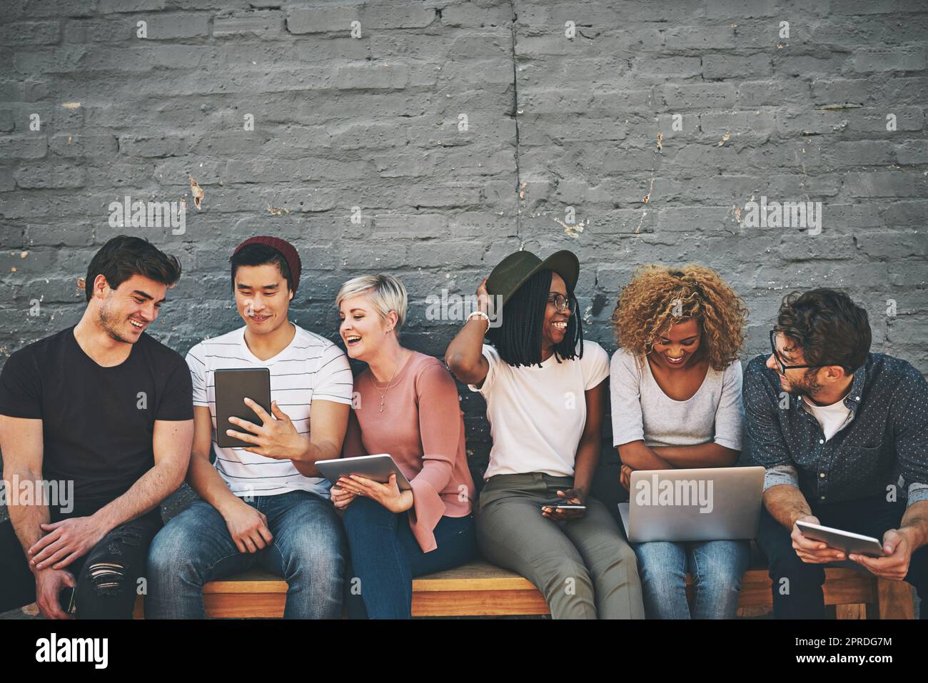 Grupo de amigos en dispositivos digitales y redes sociales sentados juntos mientras sonríen y hablan. La tecnología obsesionó a los trabajadores remotos en línea tomando un descanso. Equipo creativo casual feliz afuera Foto de stock