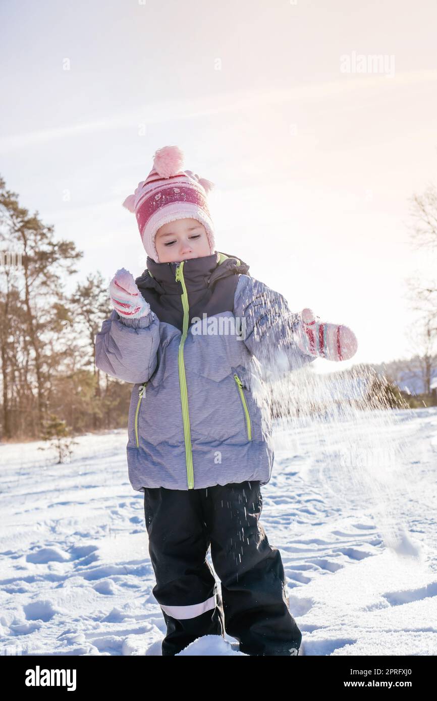 Niña de 3-4 años en el mono de invierno, sombrero y mitones, se para al  aire libre contra el cielo azul y los abetos y arroja nieve sobre su risa  Fotografía de