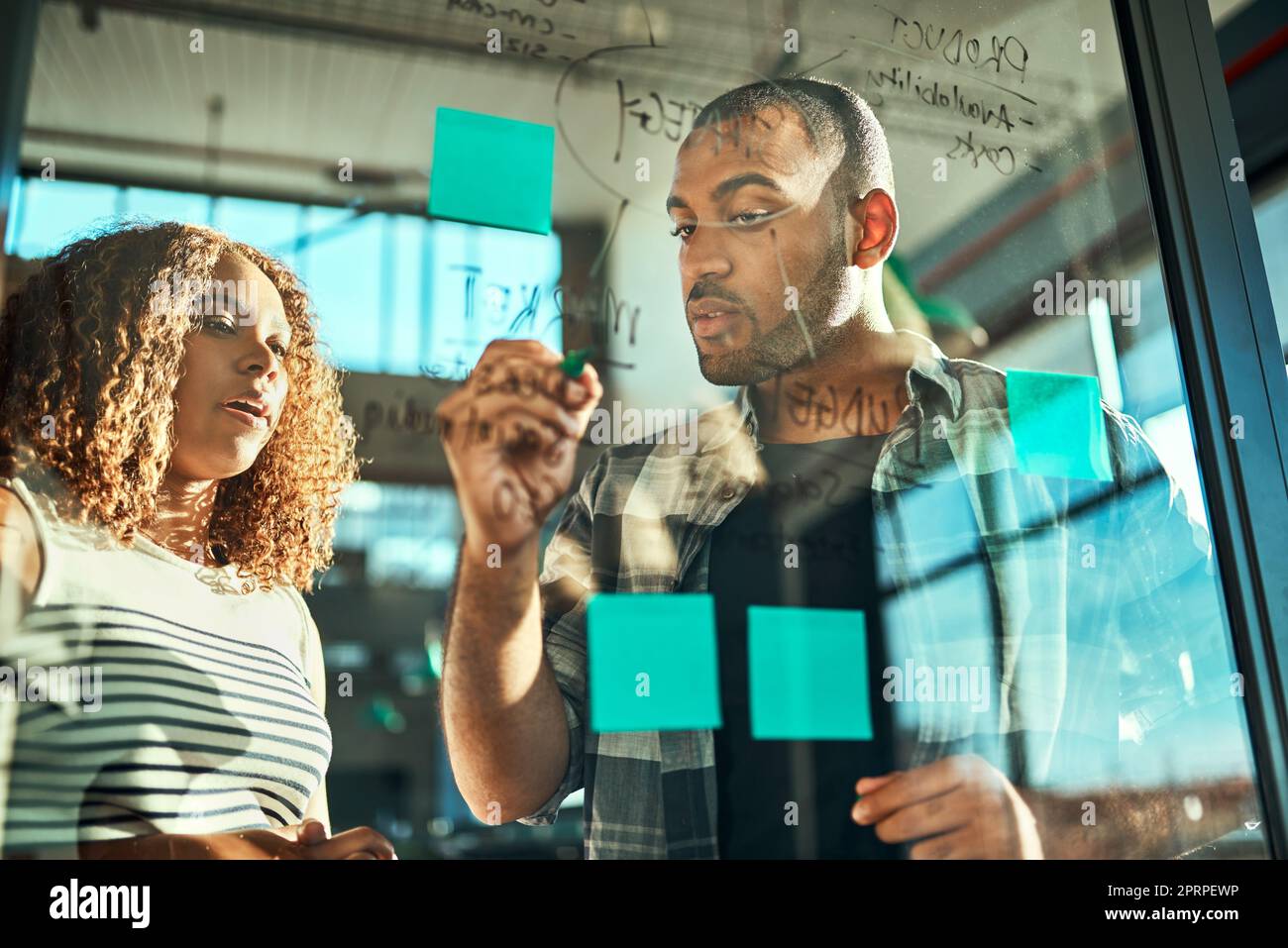 Sus mentes piensan juntas como una sola. dos jóvenes colegas de trabajo centrados que ponen notas adhesivas en una pared de vidrio mientras hacen una lluvia de ideas juntos en la oficina Foto de stock