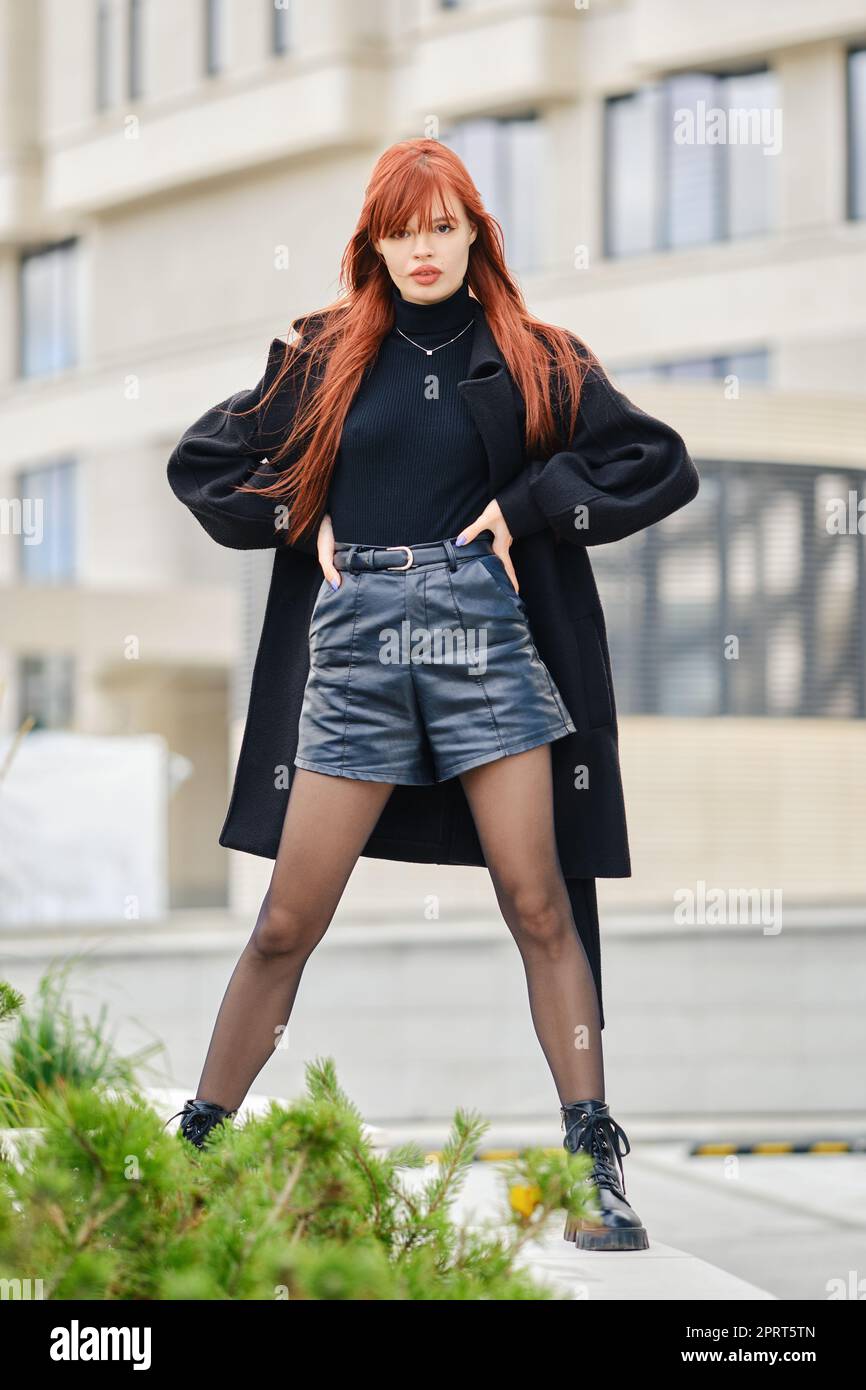 Mujer joven con estilo con pelo rojo en abrigo negro, pullover y pantalones  cortos de cuero posando en la calle Fotografía de stock - Alamy